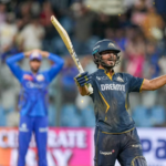 A triumphant cricketer in a dark blue and gold jersey celebrates a victory by raising his bat and clenching his fist, while a defeated opponent in blue stands blurred in the background of a crowded stadium.