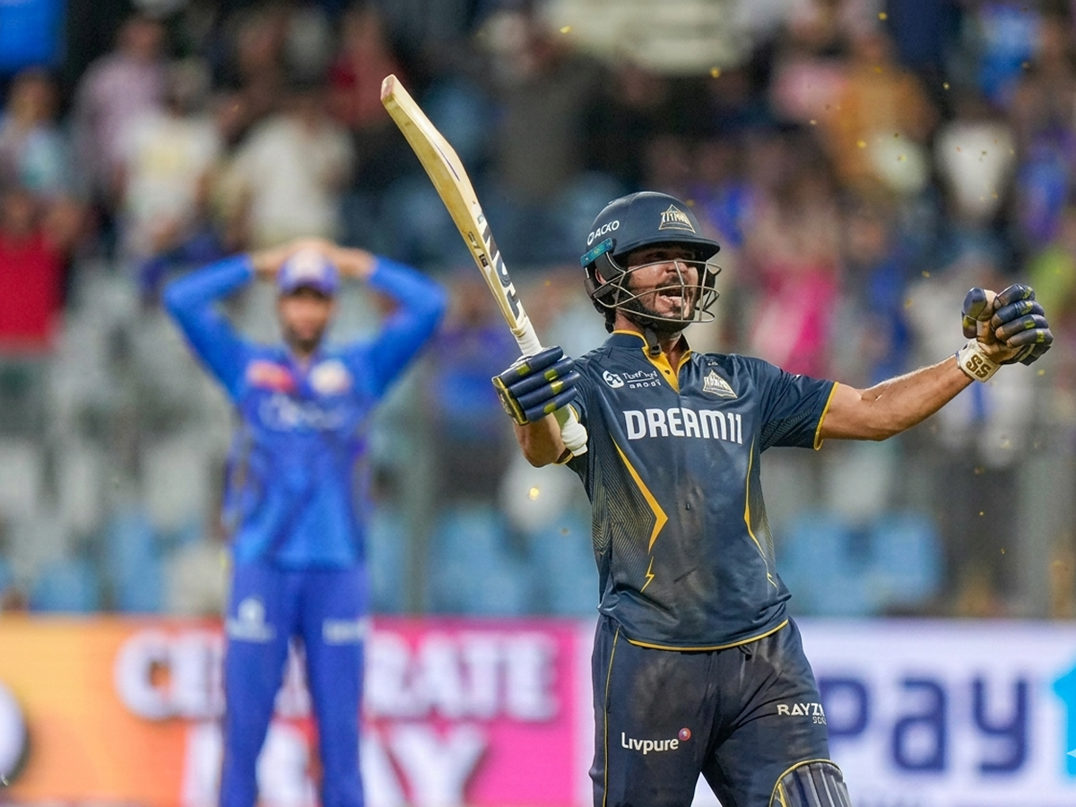 A triumphant cricketer in a dark blue and gold jersey celebrates a victory by raising his bat and clenching his fist, while a defeated opponent in blue stands blurred in the background of a crowded stadium.